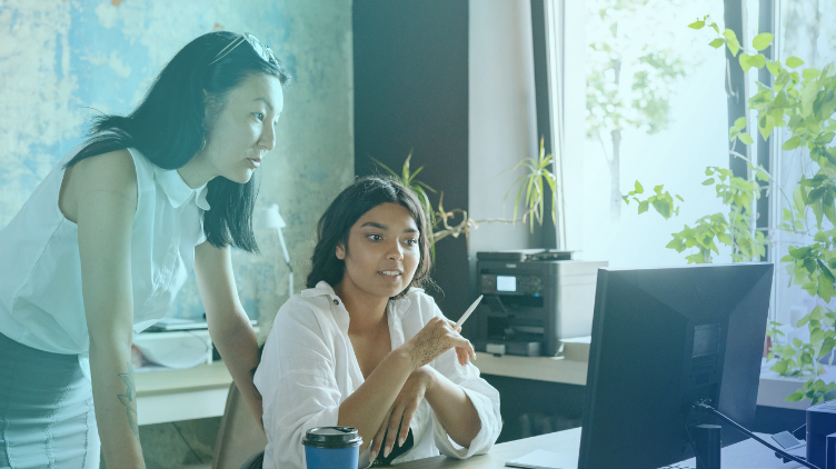 Two female professionals look at a computer screen together in a bright office filled with green plants.