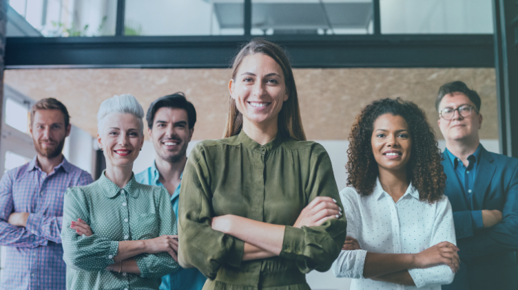 A group of six diverse, smiling business professionals with their arms crossed, standing in an office setting.