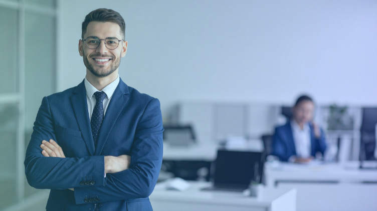 A smiling businessman in a suit standing with arms crossed in a modern office environment.